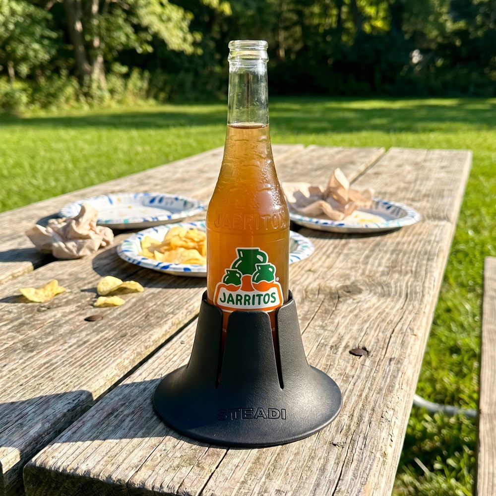 A glass soda bottle standing in a charcoal Steadi on a weathered picnic table, sunlit lawn and a wall of trees behind it.