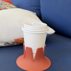 A white paper coffee cup with a lid seated in a terracotta STEADI base on the navy cushion of an outdoor patio sofa, with a linen throw pillow and palm fronds behind in bright sun