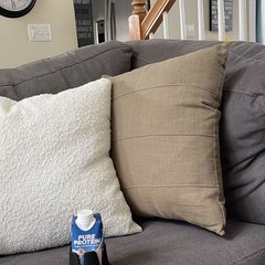 A black drink base holding a Pure Protein shake sitting on the seat cushion of a grey sectional sofa between a white boucle pillow and a tan pillow; wall clocks, staircase and kitchen behind
