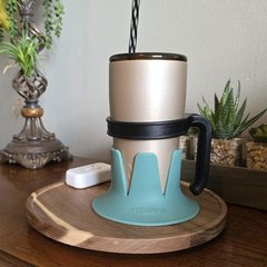 A champagne-gold tumbler with a black-and-white striped straw in a turquoise STEADI base, standing on a round wooden serving board on a dark wood side table with a lamp, a small remote and potted succulents