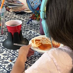 A young girl in mint headphones eating a plate of food at a kitchen table with a blue-and-white tiled tablecloth; a black drink base holds a red plastic party cup beside her plate, a birthday balloon behind
