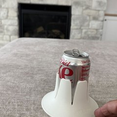 A Diet Dr Pepper can in a white drink base standing on a soft taupe blanket, a hand steadying nothing beside it, with a stone fireplace and a wall-mounted TV in the background