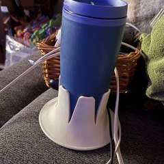 A blue travel tumbler in a cream drink base perched on the upholstered arm of a grey couch, charging cables and a folded green towel around it, dim room at night