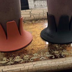 Two drink bases — coral and black — each holding a tall glass of purple berry smoothie, standing on an ornate gold-patterned serving tray in a living room with a patterned armchair behind