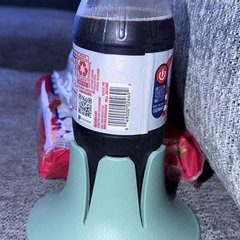 A plastic cola bottle standing in a mint-green drink base on a grey woven fabric couch cushion, wedged beside the arm